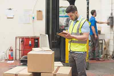 man in a high viz jacket writing on a clipboard, he is standing infront of some cardboard boxes