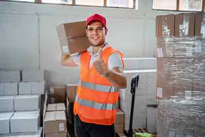 worker with a cardboard box on his shoulder giving a thumb up, he is wearing a yellow hi viz vest