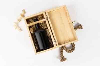 a wooden box opened on a table and there is a dark coloured bottle in it, it has a rope handle on the box
