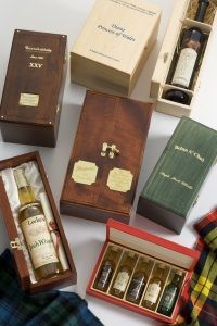 Elegant whisky collection displayed in various stylish wooden boxes, each featuring a unique bottle. Plaid fabric adds a cozy, sophisticated touch.